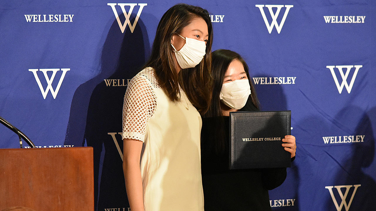 两名学生合影，其中一名学生举着她的毕业证盒，上面写着韦尔斯利学院。亚博电竞官网