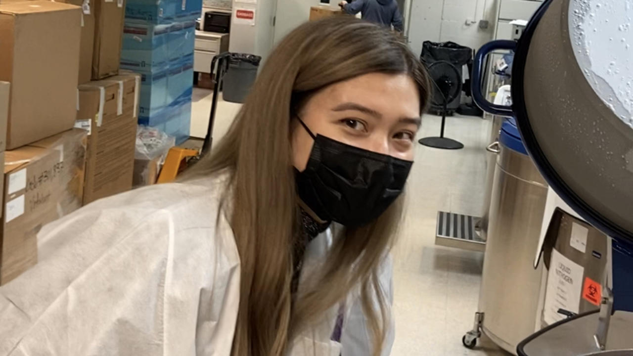 凯瑟琳穿着实验室外套和黑色面膜的交叉看着相机。
